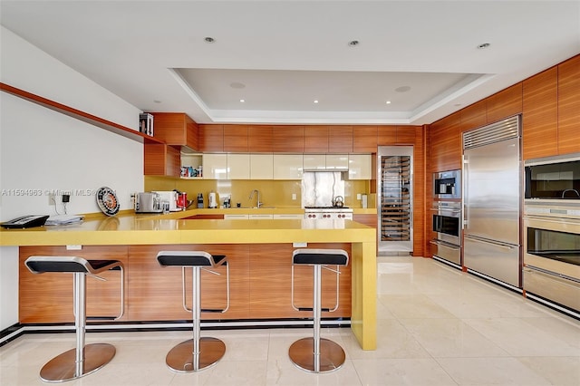 kitchen featuring a tray ceiling, built in appliances, light tile floors, sink, and a breakfast bar area