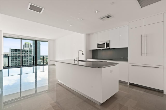 kitchen with light tile patterned flooring, white cabinets, a wall of windows, sink, and a center island with sink