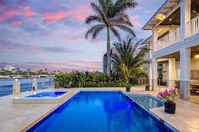 pool at dusk with an in ground hot tub, a water view, and a patio