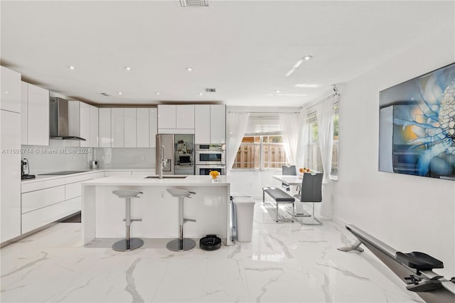 kitchen with a center island with sink, stainless steel appliances, wall chimney exhaust hood, a kitchen bar, and light tile floors