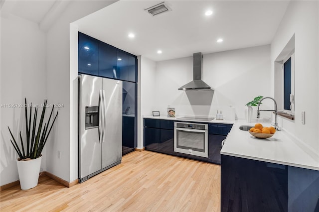 kitchen with light wood-type flooring, sink, wall chimney range hood, stainless steel appliances, and blue cabinetry