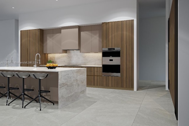 kitchen with light tile patterned floors, decorative backsplash, a kitchen island with sink, and double oven