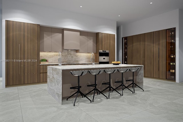 kitchen with double oven, a kitchen island with sink, light tile patterned floors, decorative backsplash, and a kitchen bar