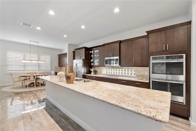 kitchen featuring backsplash, stainless steel appliances, a kitchen island with sink, pendant lighting, and light hardwood / wood-style flooring