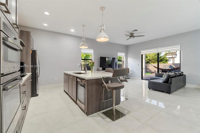 kitchen featuring ceiling fan, sink, a center island with sink, stainless steel refrigerator, and hanging light fixtures