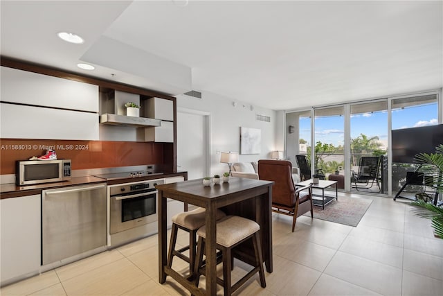 kitchen with floor to ceiling windows, stainless steel appliances, wall chimney range hood, white cabinets, and light tile patterned floors