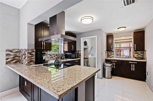 kitchen featuring washer and dryer, island exhaust hood, black appliances, backsplash, and light stone countertops