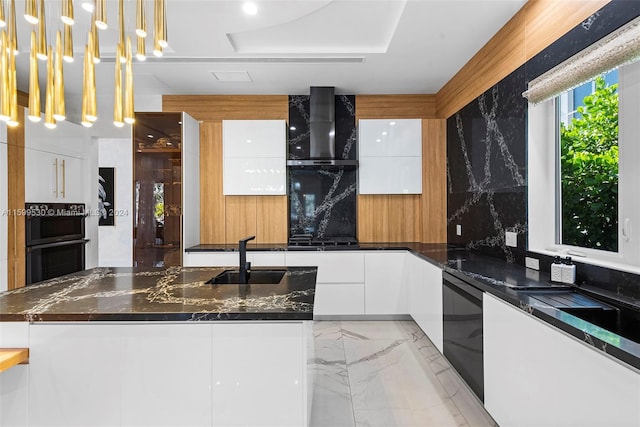 kitchen featuring wall chimney range hood, sink, dark stone countertops, decorative light fixtures, and white cabinetry