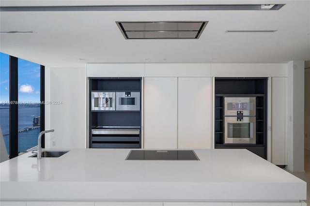 kitchen with black electric stovetop, an island with sink, white cabinets, a water view, and sink