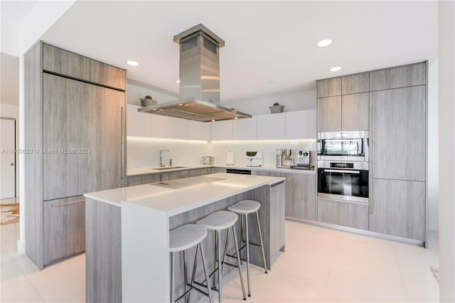 kitchen featuring island range hood, light tile flooring, a center island, a breakfast bar, and double oven