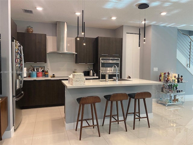 kitchen featuring wall chimney exhaust hood, stainless steel appliances, hanging light fixtures, a center island with sink, and dark brown cabinets