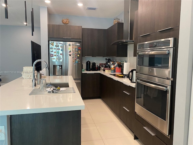 kitchen with wall chimney exhaust hood, stainless steel appliances, a kitchen island with sink, dark brown cabinets, and light tile patterned floors