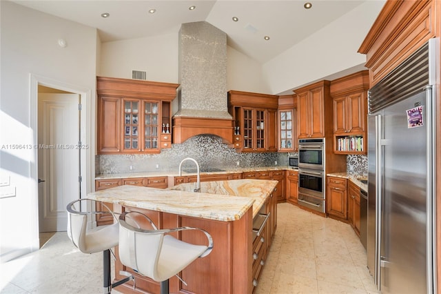 kitchen with a kitchen island with sink, stainless steel appliances, light stone countertops, backsplash, and custom exhaust hood