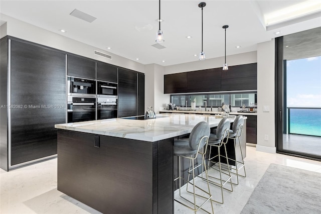 kitchen featuring light stone countertops, a breakfast bar area, double oven, pendant lighting, and a large island with sink