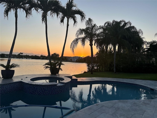 pool at dusk with an in ground hot tub, a yard, and a water view