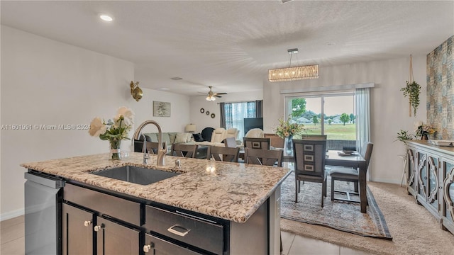 kitchen featuring ceiling fan with notable chandelier, light stone countertops, dishwasher, an island with sink, and sink