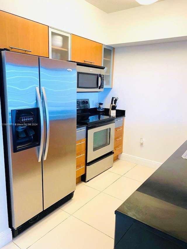 kitchen with glass insert cabinets, light tile patterned flooring, dark countertops, stainless steel appliances, and brown cabinets