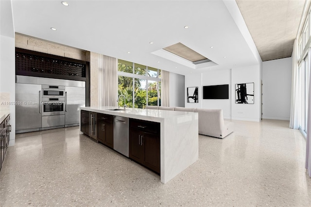 kitchen featuring a kitchen island with sink, a raised ceiling, sink, appliances with stainless steel finishes, and dark brown cabinets