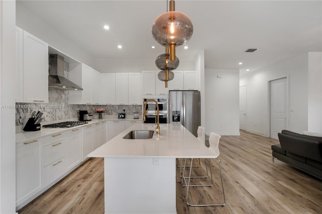 kitchen with wall chimney range hood, appliances with stainless steel finishes, decorative light fixtures, light hardwood / wood-style flooring, and a center island with sink