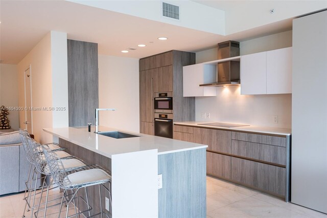 kitchen with white cabinets, wall chimney exhaust hood, stainless steel double oven, a kitchen bar, and sink