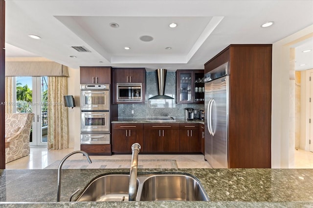 kitchen featuring a tray ceiling, built in appliances, backsplash, wall chimney exhaust hood, and sink