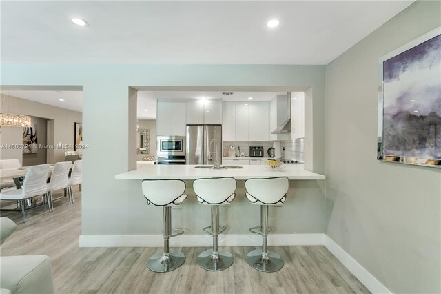 kitchen with stainless steel appliances, light hardwood / wood-style floors, white cabinetry, wall chimney range hood, and a breakfast bar area