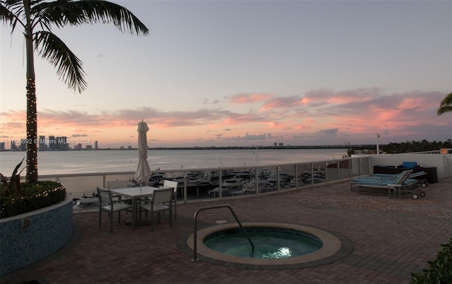 pool at dusk with a water view, a hot tub, and a patio area