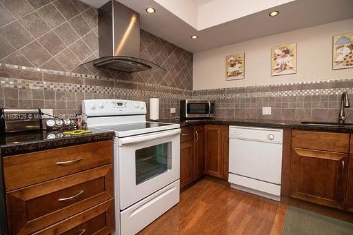 kitchen with wall chimney range hood, dark wood-type flooring, white appliances, sink, and tasteful backsplash