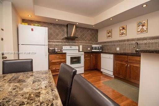 kitchen featuring white appliances, backsplash, wood-type flooring, and wall chimney range hood