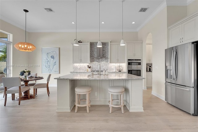 kitchen featuring wall chimney range hood, white cabinetry, hanging light fixtures, stainless steel appliances, and a center island with sink