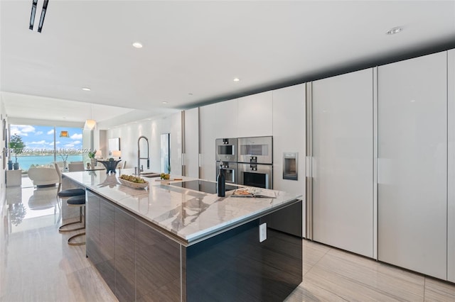 kitchen featuring cooktop, white cabinets, light stone counters, decorative light fixtures, and an island with sink