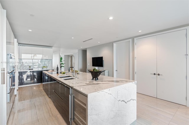 kitchen featuring light tile patterned floors, wine cooler, white cabinets, a kitchen island with sink, and light stone countertops