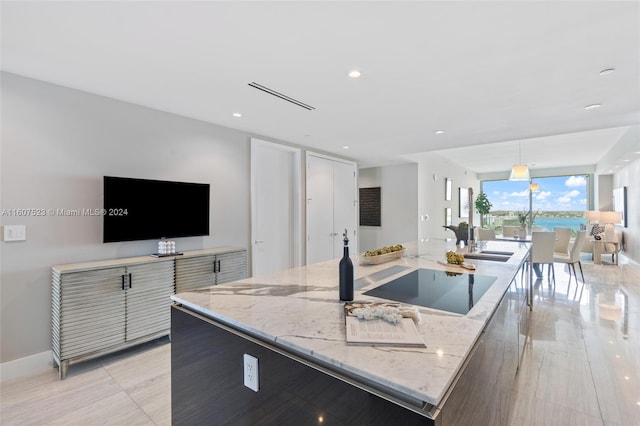 kitchen featuring light stone counters, black electric stovetop, decorative light fixtures, and an island with sink