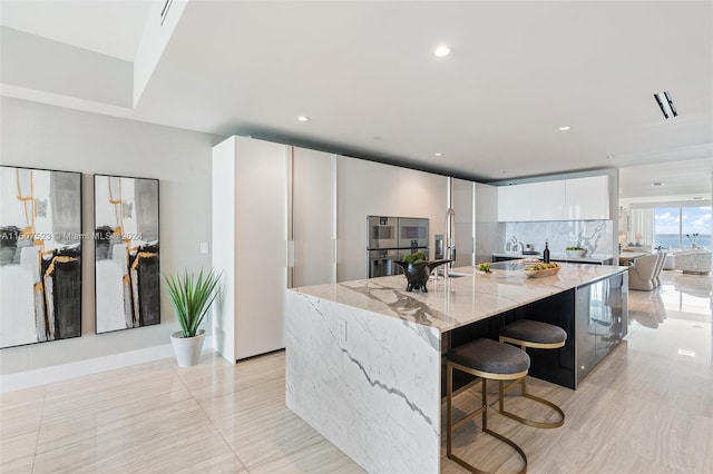 kitchen featuring white cabinets, light stone countertops, multiple ovens, a breakfast bar, and a kitchen island with sink