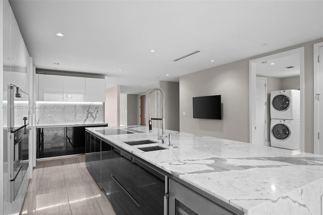 kitchen with sink, light stone countertops, stacked washer / drying machine, white cabinetry, and black electric stovetop