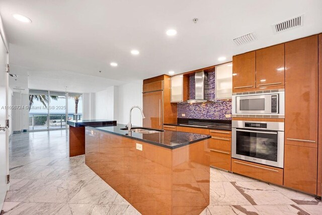 kitchen with sink, wall chimney exhaust hood, tasteful backsplash, built in appliances, and an island with sink