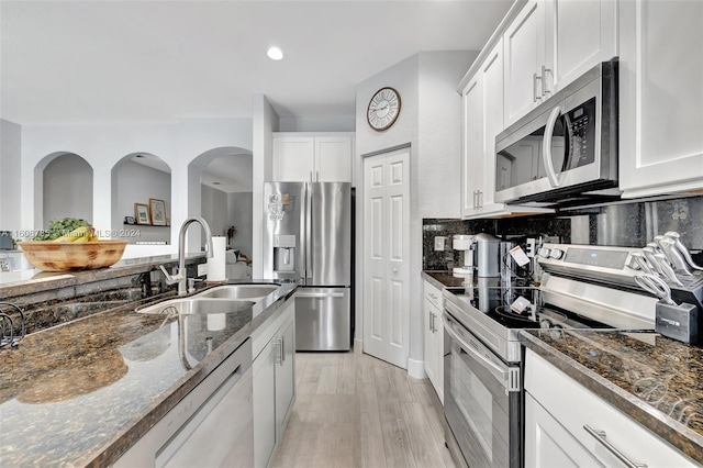 kitchen with appliances with stainless steel finishes, white cabinets, dark stone counters, light hardwood / wood-style flooring, and sink