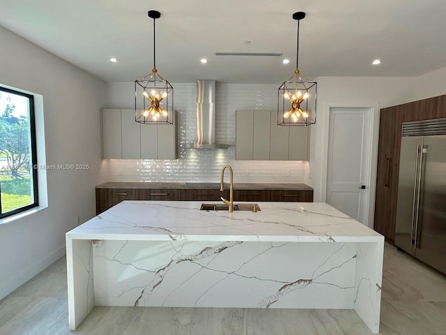 kitchen with pendant lighting, wall chimney exhaust hood, built in fridge, and an island with sink