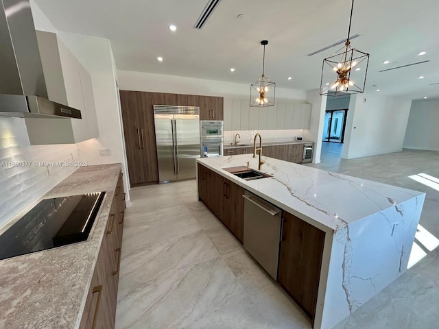 kitchen with a large island with sink, wall chimney range hood, hanging light fixtures, sink, and stainless steel appliances