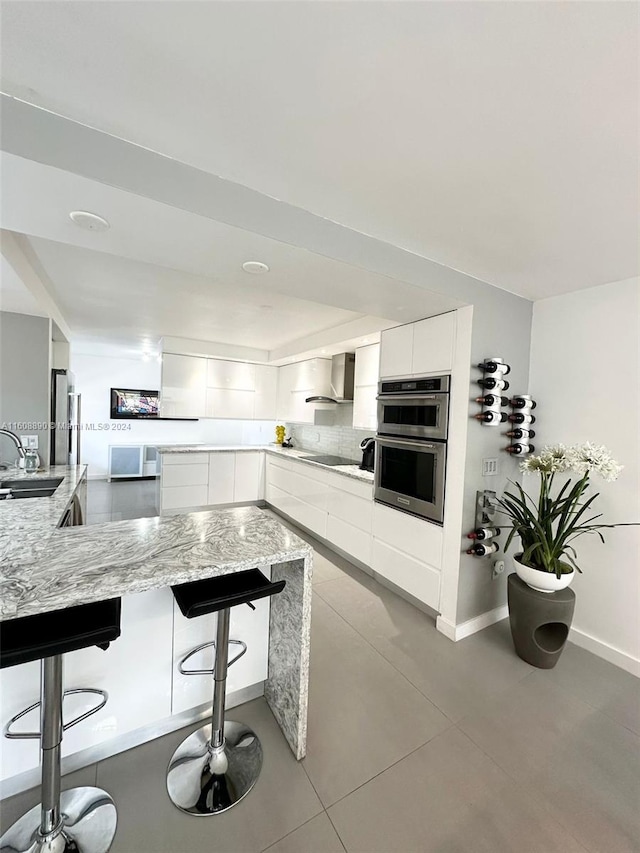 kitchen featuring white cabinetry, wall chimney exhaust hood, double oven, light stone countertops, and a breakfast bar area