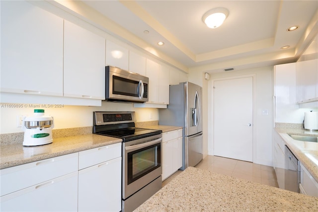 kitchen with stainless steel appliances, a tray ceiling, light tile patterned flooring, white cabinetry, and light stone counters