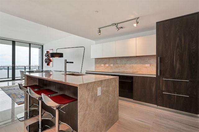 kitchen with a kitchen island with sink, black appliances, tasteful backsplash, white cabinetry, and a wall of windows