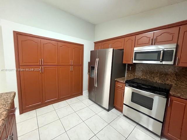 kitchen with brown cabinets, stainless steel appliances, backsplash, light tile patterned flooring, and dark stone countertops