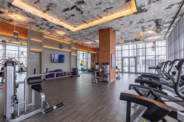 gym featuring dark hardwood / wood-style floors, ceiling fan, a tray ceiling, a wall of windows, and brick wall