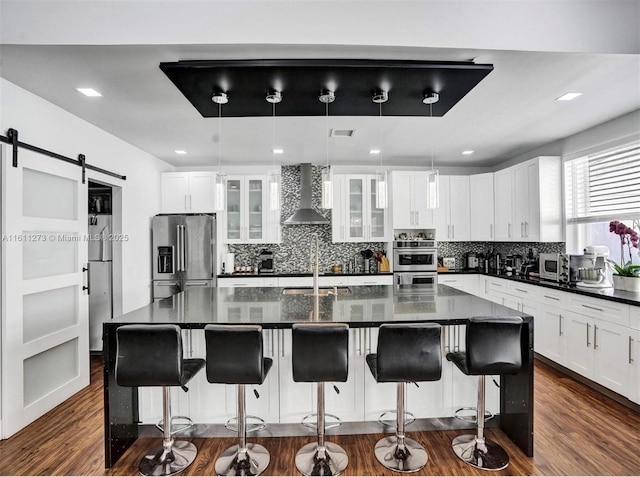 kitchen with a barn door, dark wood-type flooring, appliances with stainless steel finishes, decorative backsplash, and wall chimney exhaust hood