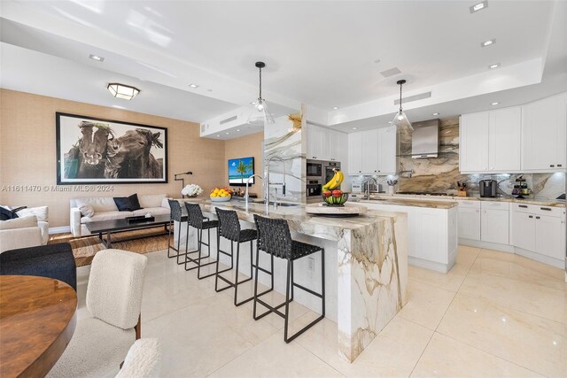 kitchen featuring tasteful backsplash, a center island with sink, wall chimney range hood, hanging light fixtures, and white cabinetry