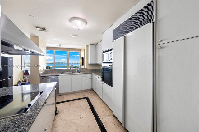 kitchen featuring white cabinetry, dark stone counters, built in appliances, wall chimney exhaust hood, and sink