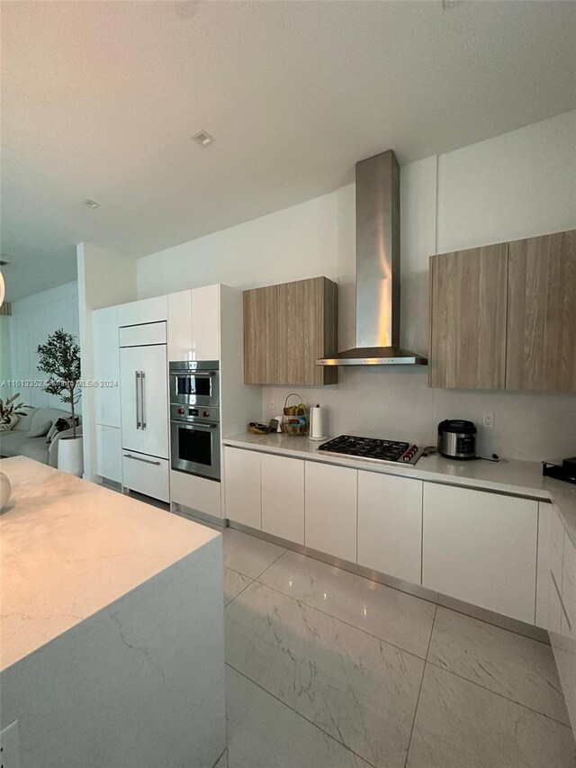 kitchen featuring gas stovetop, wall chimney range hood, paneled built in refrigerator, stainless steel double oven, and white cabinetry