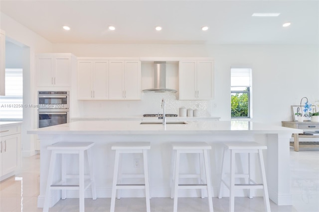 kitchen featuring wall chimney exhaust hood, white cabinetry, a kitchen island with sink, and appliances with stainless steel finishes