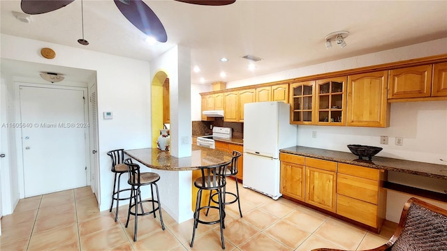 kitchen with dark stone countertops, a kitchen bar, white appliances, and light tile flooring